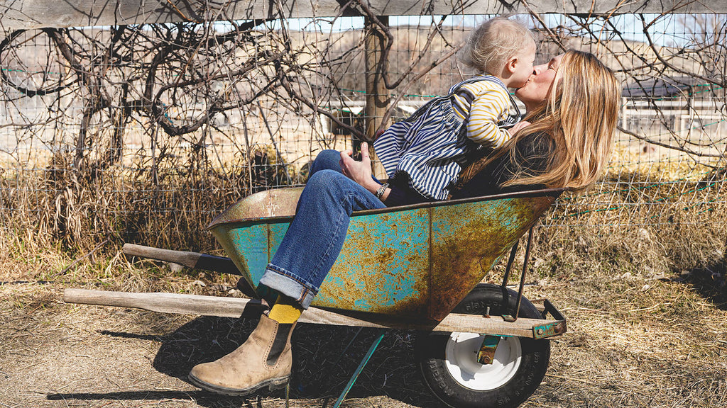 A woman with long blond hair sitting in a wheelbarrow kissing her young daughter who is sitting in her lap.