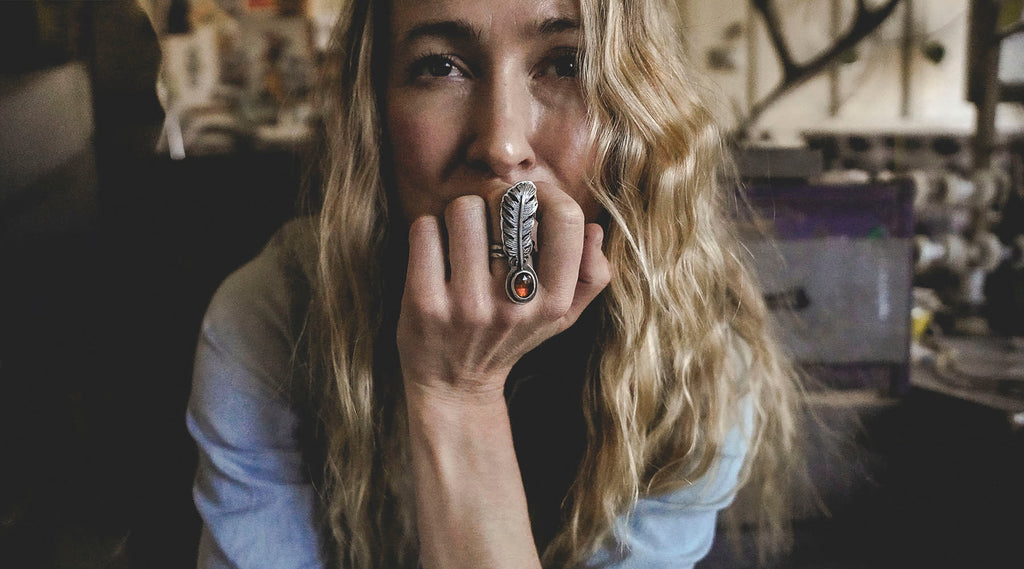 A woman with long blonde hair looking directly into the camera. She is wearing a blue chambray shirt and a sterling silver and hessonite ring. The ring has a feather shape on the front and the gemstone is dark orange-red. 