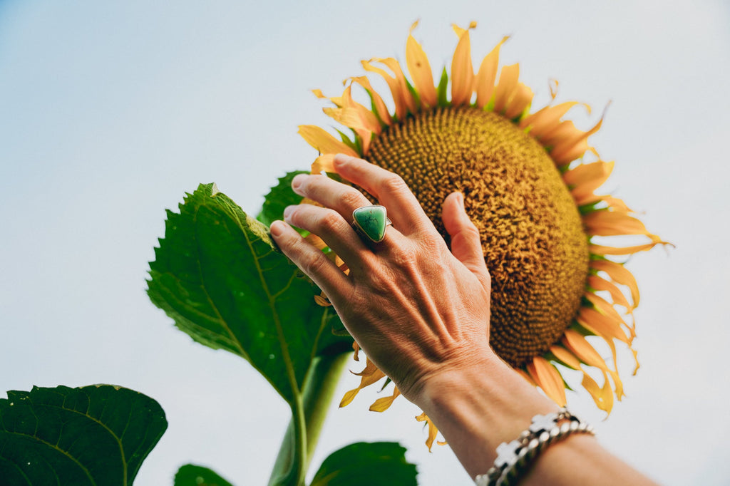 A woman's hand against a large sunflower. She ie wearing a chucnky silver and turquoise ring and sulver bracelet with c=numerous connected crosses.