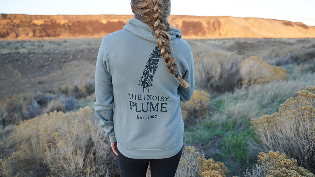 A woman with long blonde hair in a braid stands on a high desert plateau. She is wearing a blue-gray hooded sweatshirt that has a feather and the words "The Noisy Plume" printed on the back. There are sunlit mountains in the distance.