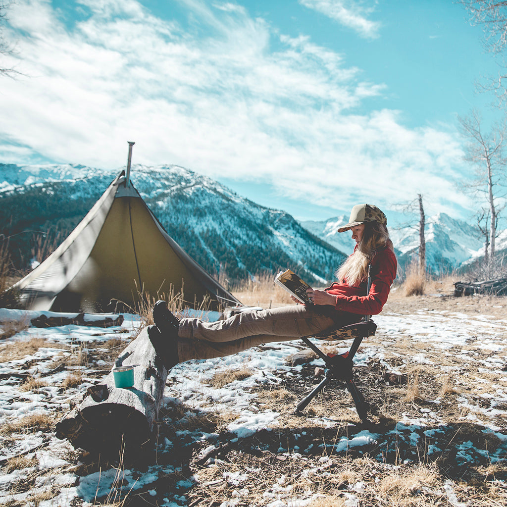 Woman sitting on a camp chair feet on a log reading a book with a tent and snowy mountains in the background