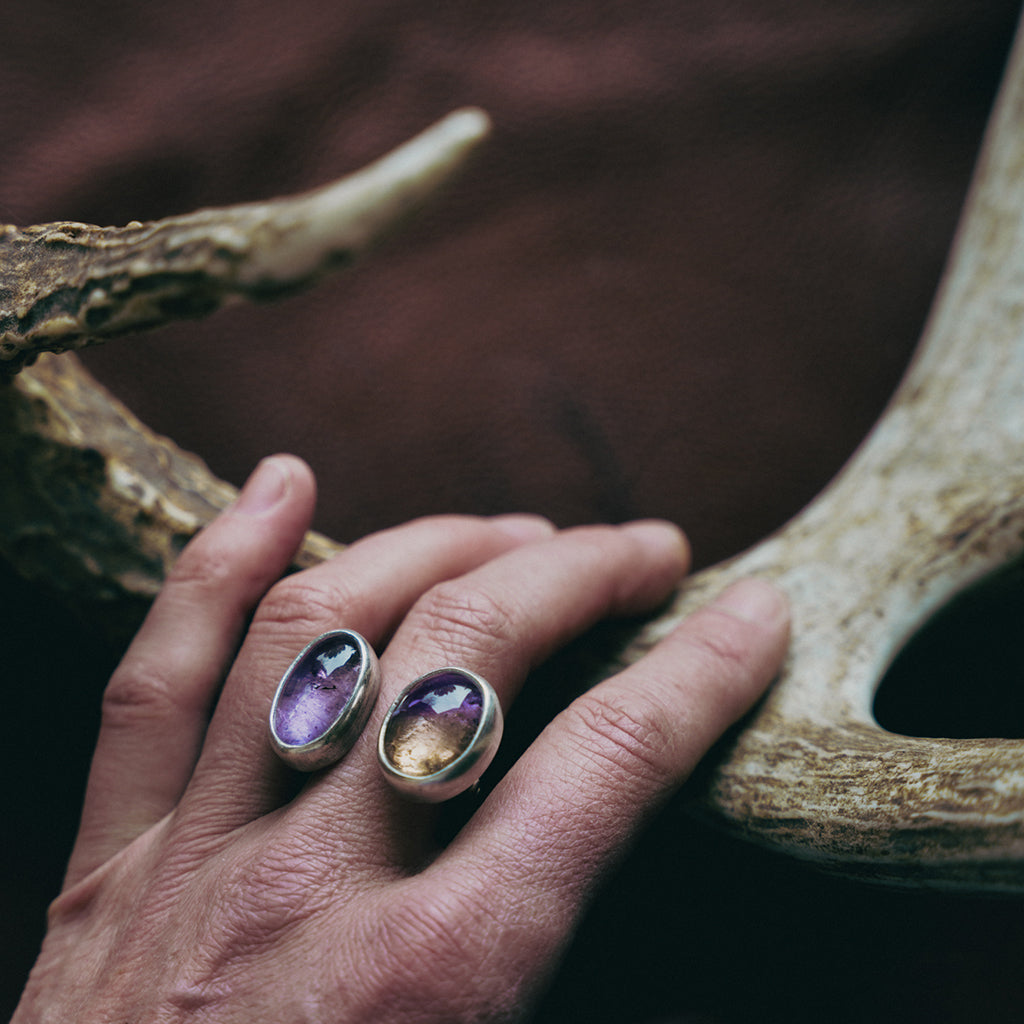 Hand wearing a chunky sterling silver double-sided ring with purple and pale yellow gemstones resting on an antler.
